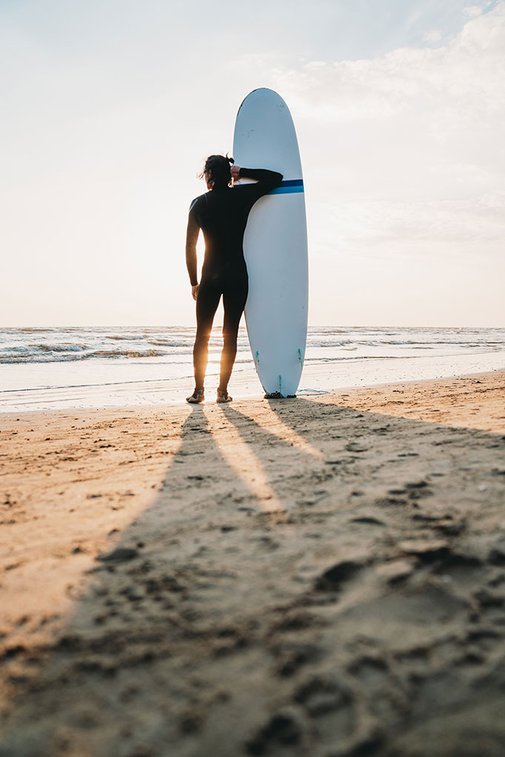 Ein Mann steht mit seinem Surfbrett am Strand und bewundert den Sonnenuntergang. Rückansicht von ihm mit Blick auf das Meer symbolisiert Hotels in Corralejo